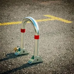 Metalowa blokada na miejsce parkingowe, zabezpieczona kłódką, stoi na asfalcie obok żółtej linii.