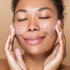 Kobieta z zamkniętymi oczami delikatnie wmasowuje peeling do twarzy. Jej skóra promienieje po zabiegu.