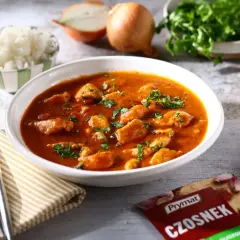 Aksamitny gulasz z piersi kurczaka w pomidorowym sosie, posypany natką pietruszki. Obok miseczka ryżu, cebula i przyprawa czosnkowa.