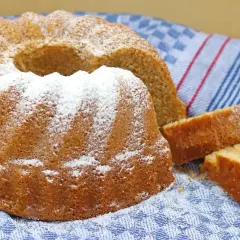 Puszysta babka na oleju, oprószona cukrem pudrem, leży na niebiesko-białym ręczniku. Obok niej leżą dwa kawałki ciasta.