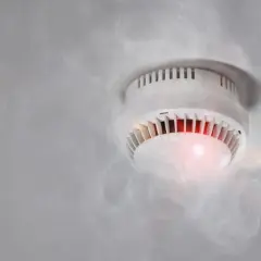 Czujnik czadu pika, otoczony kłębami dymu. Czerwona dioda sygnalizuje alarm.