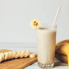 Gęsty koktajl bananowy w szklance z plasterkiem banana, obok pokrojone banany i całe owoce.