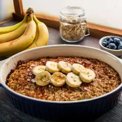 Pyszna, pieczona owsianka z bananem, udekorowana plasterkami owocu i posypana cynamonem. Obok miseczka z jagodami i pęczek bananów.