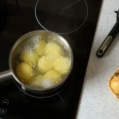 Ziemniaki gotują się w garnku na płycie indukcyjnej. Obok leży obieraczka i koszyk z surowymi ziemniakami. Czas gotowania zależy od wielkości.