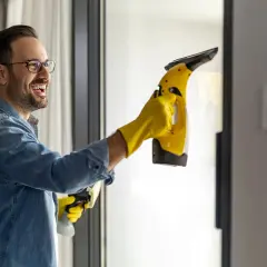 Uśmiechnięty mężczyzna w żółtych rękawiczkach pokazuje, jak myć okna ściągaczką, używając elektrycznego urządzenia.