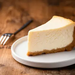 Kawałek klasycznego new york cheesecake na białym talerzyku, obok widelec, na drewnianym tle.