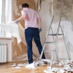 Mężczyzna w rękawiczkach zrywa starą tapetę ze ściany. Obok stoi drabina, a podłoga jest zasypana kawałkami papieru. To dowód na to, jak zerwać tapetę.