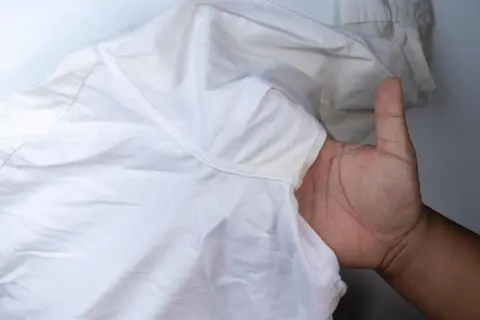 Dłoń pokazuje białą koszulę z żółtymi, twardymi plamami po antyperspirancie pod pachą.
