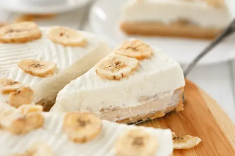 Kawałki ciasta bananowego bez pieczenia, ozdobione plasterkami suszonego banana, na drewnianej desce.