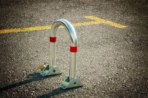 Metalowa blokada na miejsce parkingowe, zabezpieczona kłódką, stoi na asfalcie obok żółtej linii.