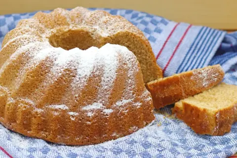 Puszysta babka na oleju, oprószona cukrem pudrem, leży na niebiesko-białym ręczniku. Obok niej leżą dwa kawałki ciasta.
