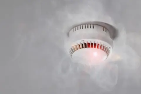 Czujnik czadu pika, otoczony kłębami dymu. Czerwona dioda sygnalizuje alarm.