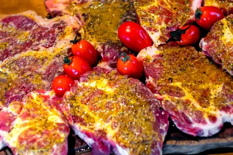 Soczyste plastry karkówki, marynowane i przyprawione, gotowe do grillowania. Obok leżą czerwone pomidorki koktajlowe.