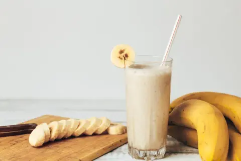 Gęsty koktajl bananowy w szklance z plasterkiem banana, obok pokrojone banany i całe owoce.