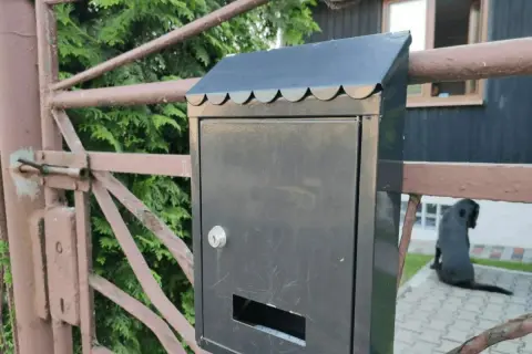 Czarna skrzynka pocztowa z nowym zamkiem, gotowa na wymianę zamka. W tle pies i dom.