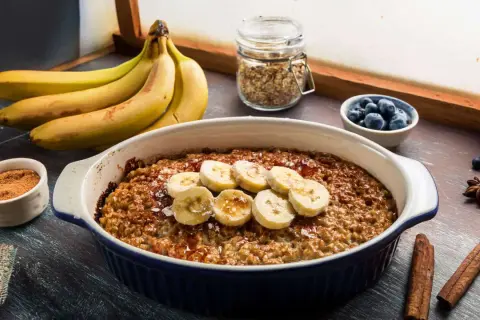 Pyszna, pieczona owsianka z bananem, udekorowana plasterkami owocu i posypana cynamonem. Obok miseczka z jagodami i pęczek bananów.