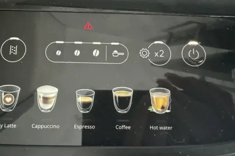 Ekspres DeLonghi z czerwoną kontrolką ostrzegawczą. Widoczne ikony napojów: latte, cappuccino, espresso, kawa, gorąca woda.