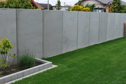 Nowy, wysoki, betonowy płot stanowi pełne ogrodzenie od sąsiada, oddzielając dwie posesje.