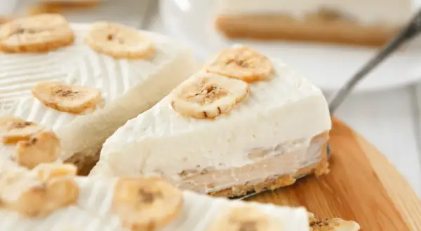 Kawałki ciasta bananowego bez pieczenia, ozdobione plasterkami suszonego banana, na drewnianej desce.