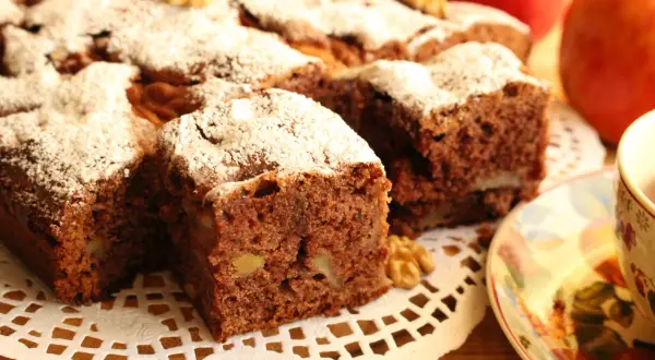 Pyszne ciasto cynamonowe z jabłkami, posypane cukrem pudrem i orzechami. Obok filiżanka herbaty i jabłka.