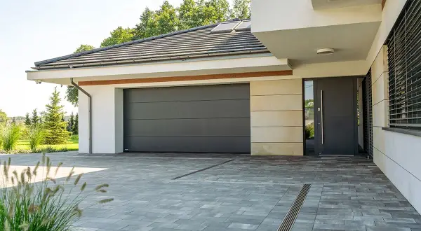 Nowoczesna brama garażowa, szara, segmentowa, z widocznymi panelami. Obok drzwi wejściowe.