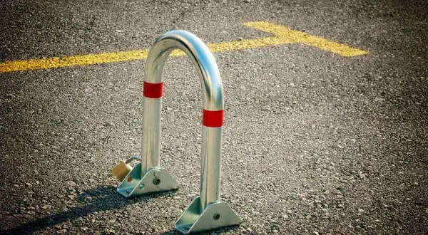 Metalowa blokada na miejsce parkingowe, zabezpieczona kłódką, stoi na asfalcie obok żółtej linii.