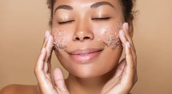 Kobieta z zamkniętymi oczami delikatnie wmasowuje peeling do twarzy. Jej skóra promienieje po zabiegu.