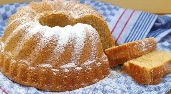 Puszysta babka na oleju, oprószona cukrem pudrem, leży na niebiesko-białym ręczniku. Obok niej leżą dwa kawałki ciasta.