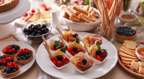 Pyszne menu na urodziny w domu: tartaletki z owocami, przekąski z szynką i oliwkami, sery, grissini i ciasto.