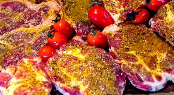 Soczyste plastry karkówki, marynowane i przyprawione, gotowe do grillowania. Obok leżą czerwone pomidorki koktajlowe.