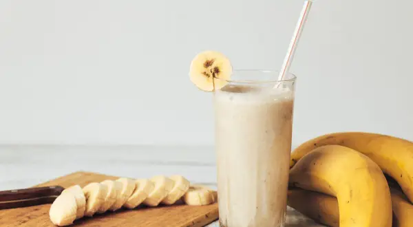 Gęsty koktajl bananowy w szklance z plasterkiem banana, obok pokrojone banany i całe owoce.
