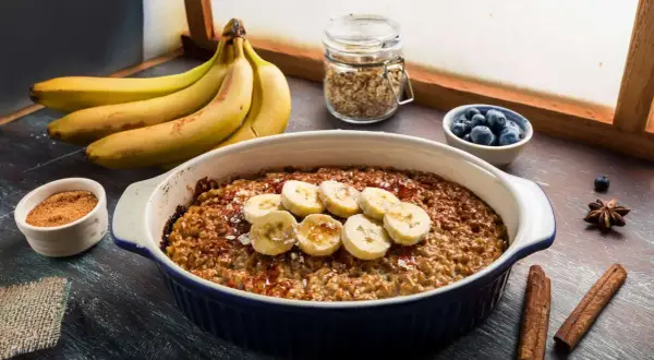 Pyszna, pieczona owsianka z bananem, udekorowana plasterkami owocu i posypana cynamonem. Obok miseczka z jagodami i pęczek bananów.