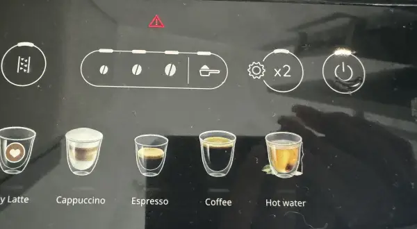 Ekspres DeLonghi z czerwoną kontrolką ostrzegawczą. Widoczne ikony napojów: latte, cappuccino, espresso, kawa, gorąca woda.