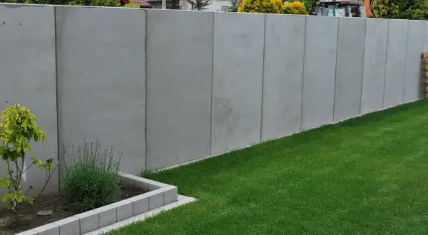 Nowy, wysoki, betonowy płot stanowi pełne ogrodzenie od sąsiada, oddzielając dwie posesje.