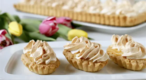 Słodkie babeczki z bezą, idealne, gdy zastanawiasz się, co można zrobić z żółtek. W tle duża tarta i tulipany.