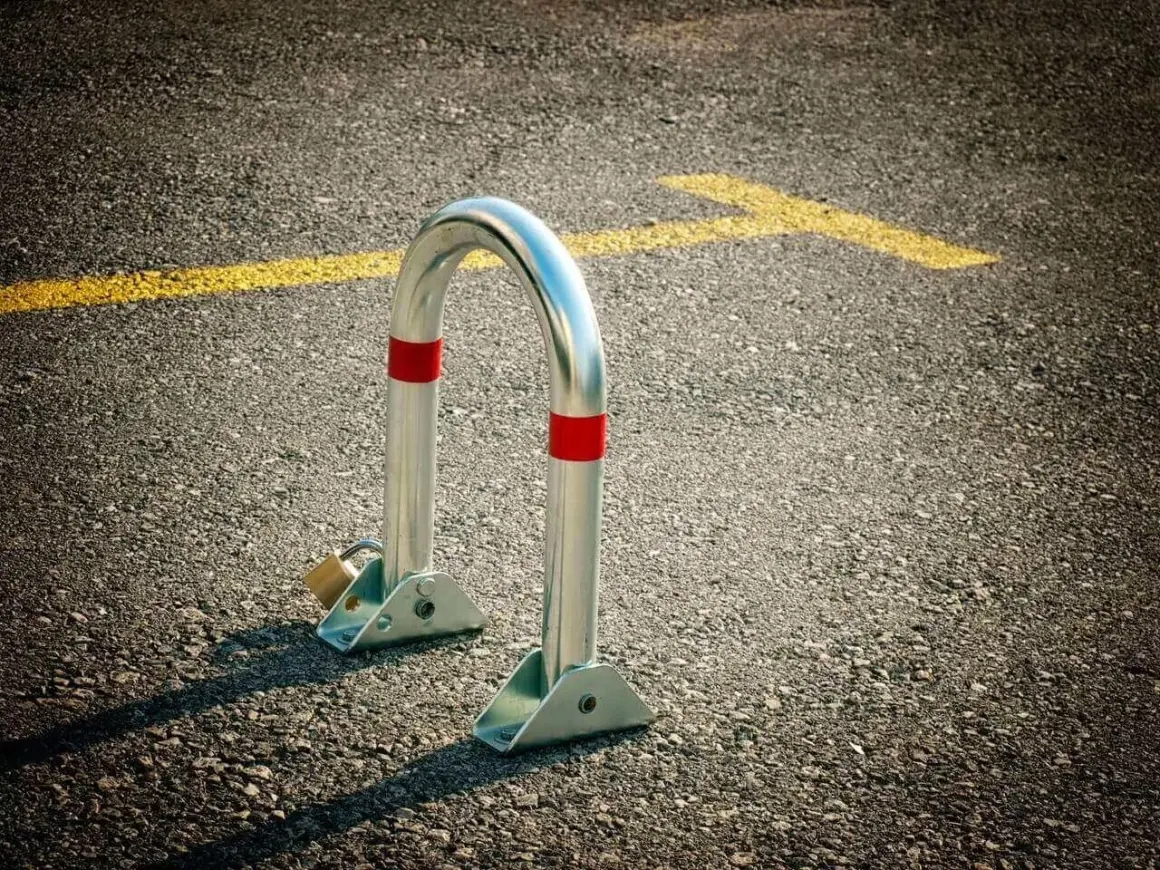 Metalowa blokada na miejsce parkingowe, zabezpieczona kłódką, stoi na asfalcie obok żółtej linii.