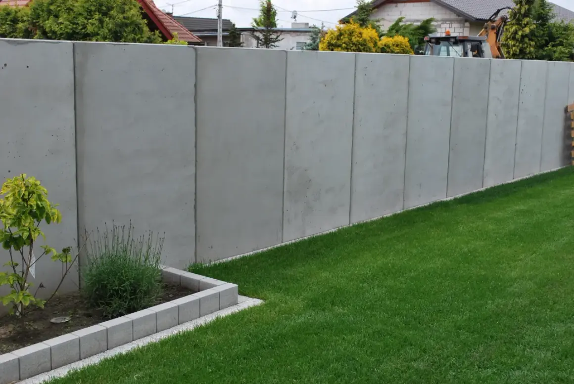 Nowy, wysoki, betonowy płot stanowi pełne ogrodzenie od sąsiada, oddzielając dwie posesje.