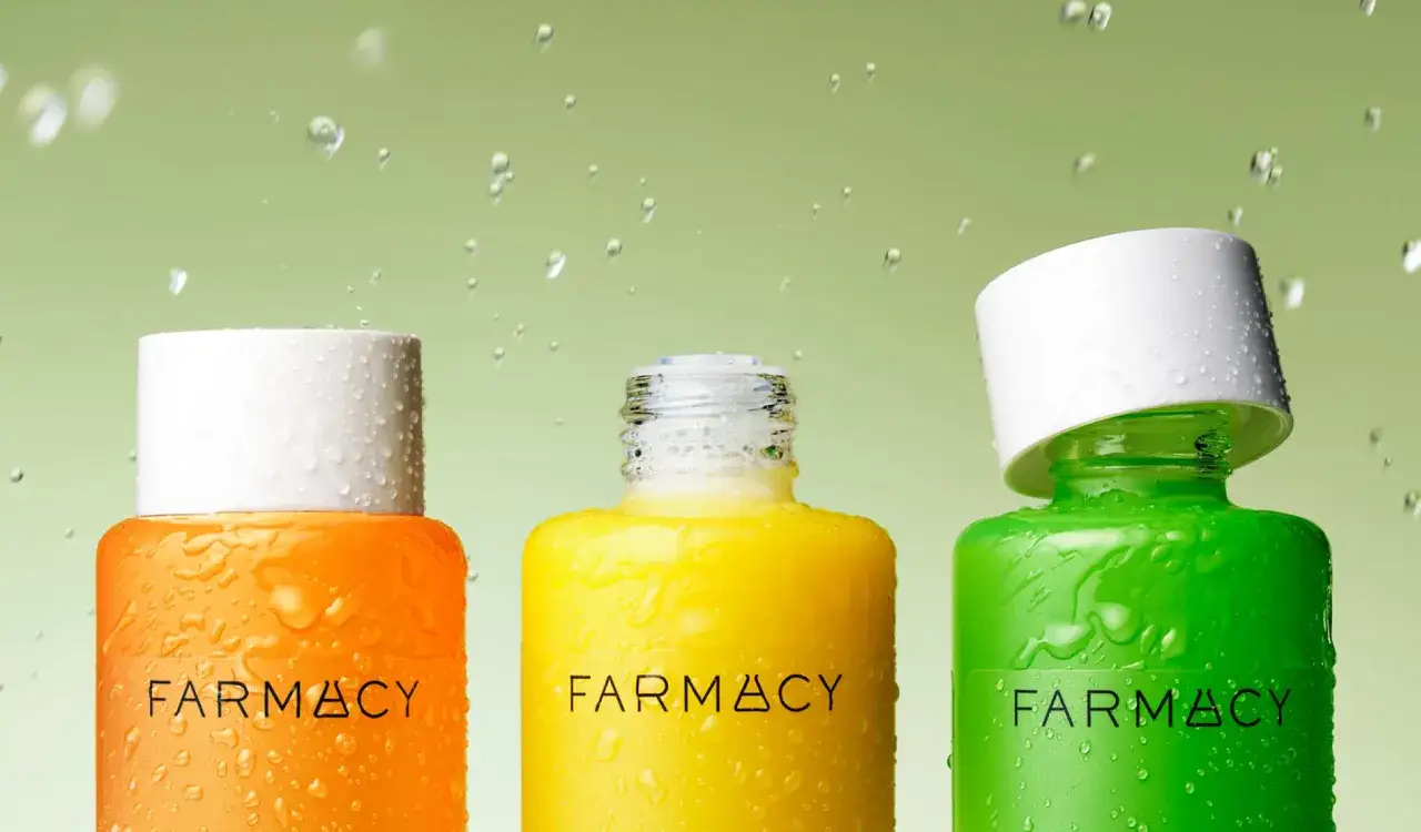 Trzy kolorowe butelki Farmacy: pomarańczowa, żółta i zielona, pokryte kroplami wody. Każda zawiera esencję do twarzy, gotową do odświeżenia skóry.