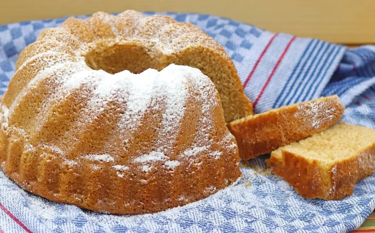 Puszysta babka na oleju, oprószona cukrem pudrem, leży obok pokrojonych kawałków na niebiesko-białym ręczniku.