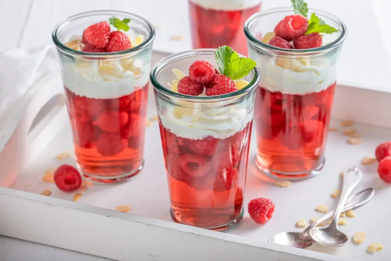 Mini deserki z galaretką malinową, bitą śmietaną i płatkami migdałów, ozdobione malinami i miętą.