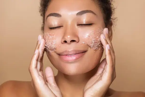 Kobieta z zamkniętymi oczami delikatnie wmasowuje peeling do twarzy. Jej skóra promienieje po zabiegu.