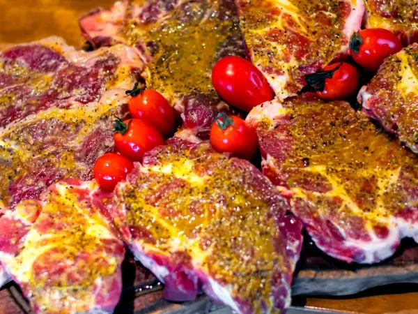 Soczyste plastry karkówki, marynowane i przyprawione, gotowe do grillowania. Obok leżą czerwone pomidorki koktajlowe.