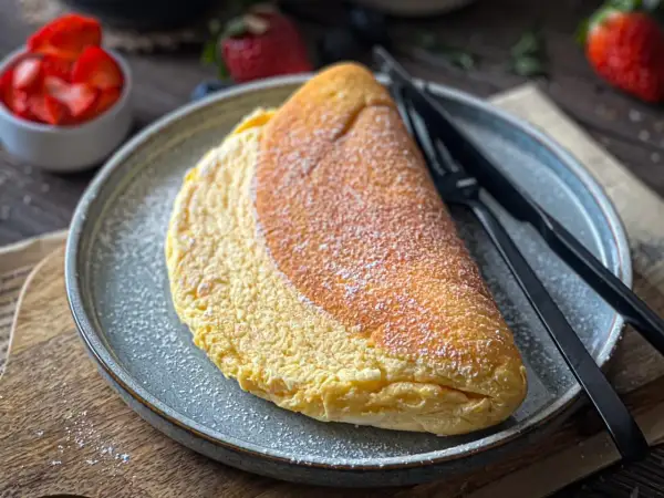 Puszysty omlet na słodko, posypany cukrem pudrem, czeka na talerzu obok widelca i noża. W tle miseczka z truskawkami.