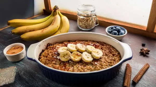 Pyszna, pieczona owsianka z bananem, udekorowana plasterkami owocu i posypana cynamonem. Obok miseczka z jagodami i pęczek bananów.