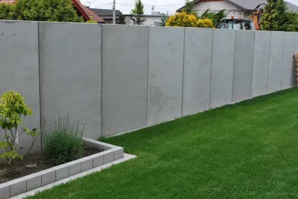 Nowy, wysoki, betonowy płot stanowi pełne ogrodzenie od sąsiada, oddzielając dwie posesje.